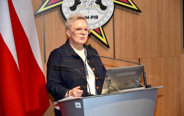 Lidia Wojda przy mównicy w sali Wojskowego Instytutu Medycznego; obok polska flaga i emblemat WIM‑PIB.