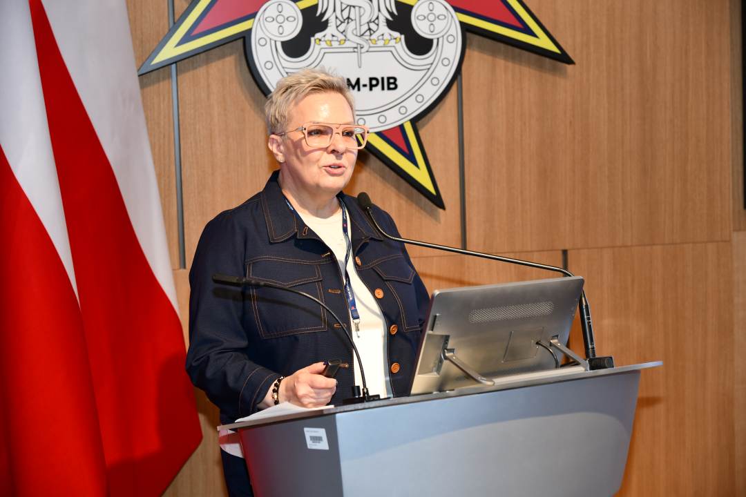 Lidia Wojda przy mównicy w sali Wojskowego Instytutu Medycznego; obok polska flaga i emblemat WIM‑PIB.