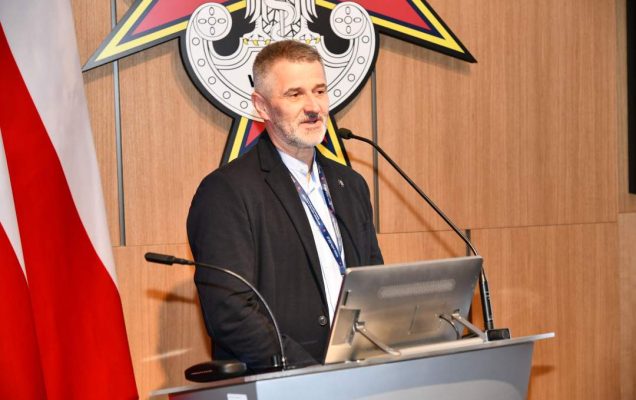 Zbigniew Wojda przy mównicy w sali Wojskowego Instytutu Medycznego; obok polska flaga i emblemat WIM‑PIB.