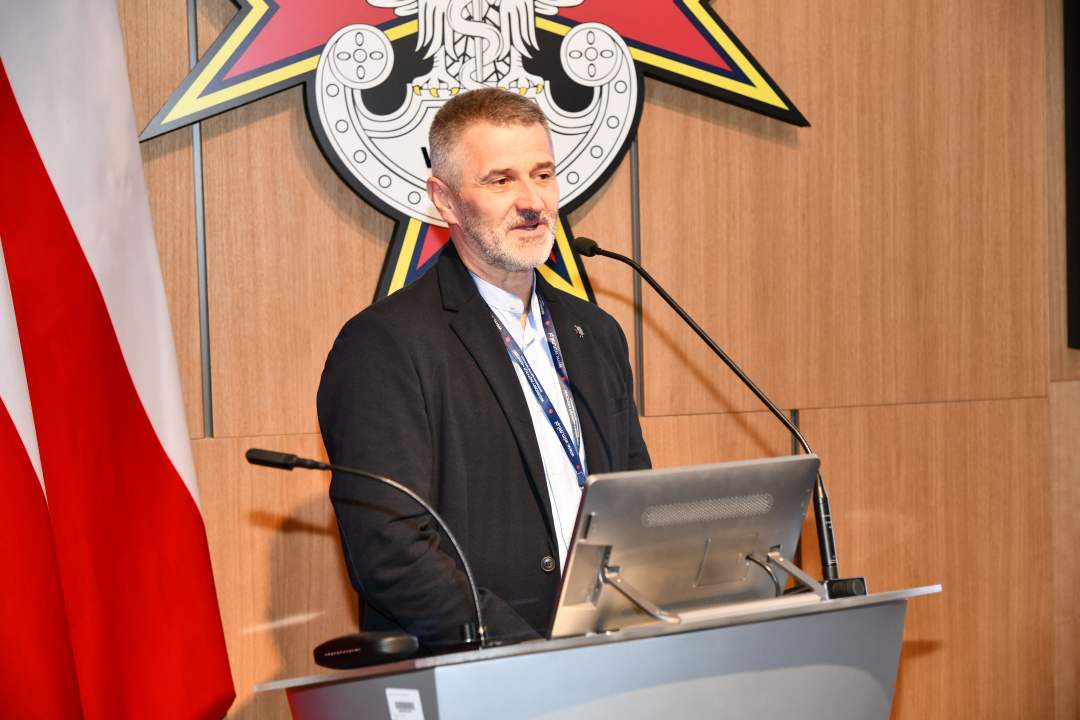 Zbigniew Wojda przy mównicy w sali Wojskowego Instytutu Medycznego; obok polska flaga i emblemat WIM‑PIB.