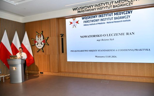 Bożena Styk przy mównicy w sali Wojskowego Instytutu Medycznego; za nią trzy polskie flagi i emblemat WIM‑PIB. Na ekranie widoczny slajd z tytułem prezentacji Nowatorsko o leczeniu ran.