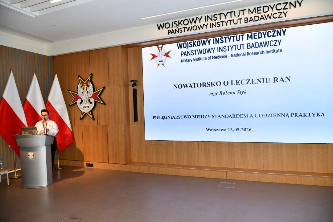 Bożena Styk przy mównicy w sali Wojskowego Instytutu Medycznego; za nią trzy polskie flagi i emblemat WIM‑PIB. Na ekranie widoczny slajd z tytułem prezentacji Nowatorsko o leczeniu ran.