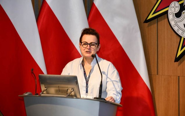 Bożena Styk przy mównicy w sali Wojskowego Instytutu Medycznego; za nią trzy polskie flagi i emblemat WIM‑PIB.