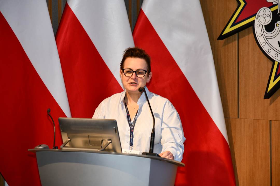 Bożena Styk przy mównicy w sali Wojskowego Instytutu Medycznego; za nią trzy polskie flagi i emblemat WIM‑PIB.