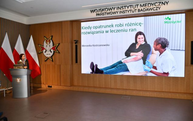 Bożena Styk przy mównicy w sali Wojskowego Instytutu Medycznego; za nią trzy polskie flagi i emblemat WIM‑PIB. Na ekranie widoczny slajd z tytułem prezentacji ‘Kiedy opatrunek robi różnicę – rozwiązania w leczeniu ran’ oraz logo Mölnlycke.