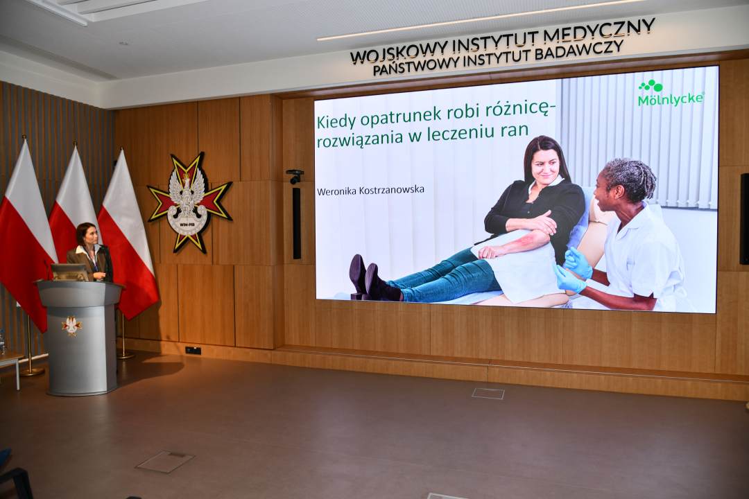 Bożena Styk przy mównicy w sali Wojskowego Instytutu Medycznego; za nią trzy polskie flagi i emblemat WIM‑PIB. Na ekranie widoczny slajd z tytułem prezentacji ‘Kiedy opatrunek robi różnicę – rozwiązania w leczeniu ran’ oraz logo Mölnlycke.