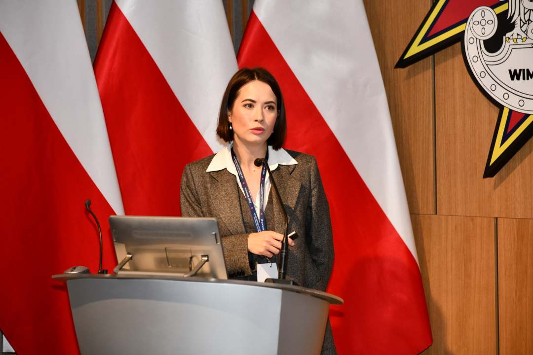 Bożena Styk przy mównicy w sali Wojskowego Instytutu Medycznego; za nią trzy polskie flagi i emblemat WIM‑PIB.