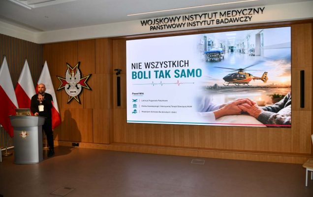 Paweł Witt przy mównicy w sali Wojskowego Instytutu Medycznego; za nim trzy polskie flagi i emblemat WIM‑PIB. Na ekranie widoczny slajd z tytułem prezentacji ‘Nie wszystkich boli tak samo’ oraz grafikami medycznymi.