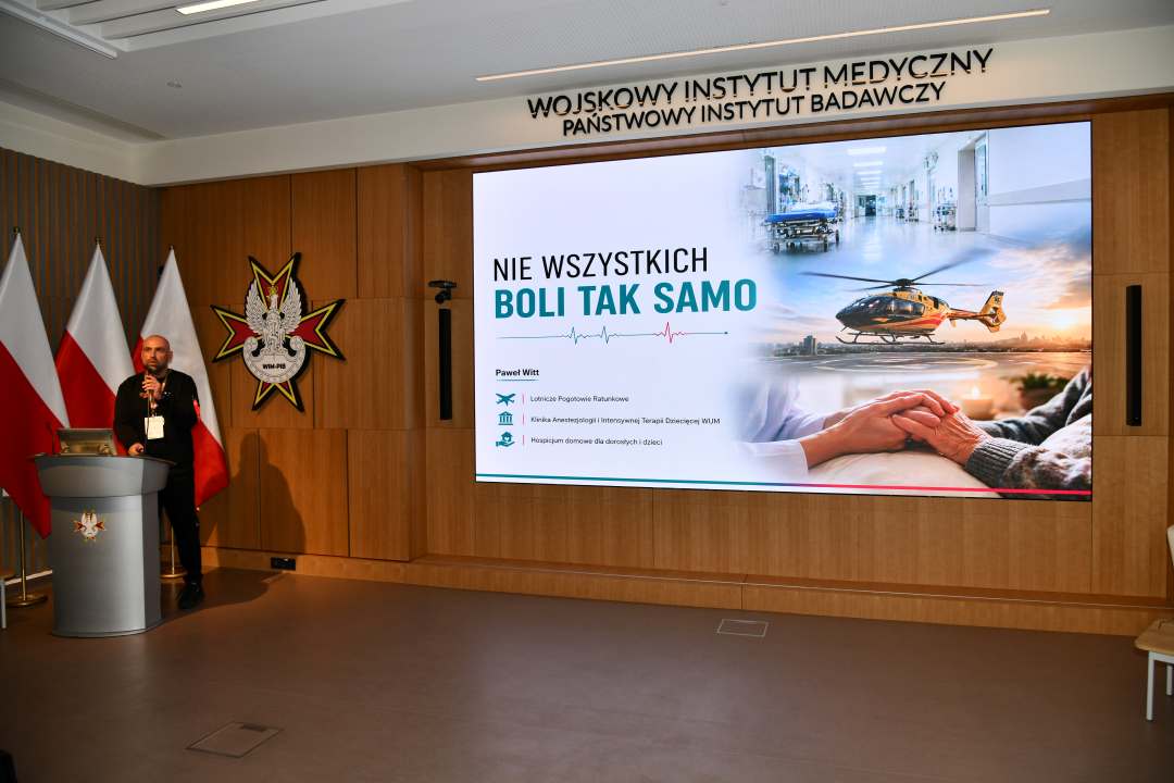 Paweł Witt przy mównicy w sali Wojskowego Instytutu Medycznego; za nim trzy polskie flagi i emblemat WIM‑PIB. Na ekranie widoczny slajd z tytułem prezentacji ‘Nie wszystkich boli tak samo’ oraz grafikami medycznymi.