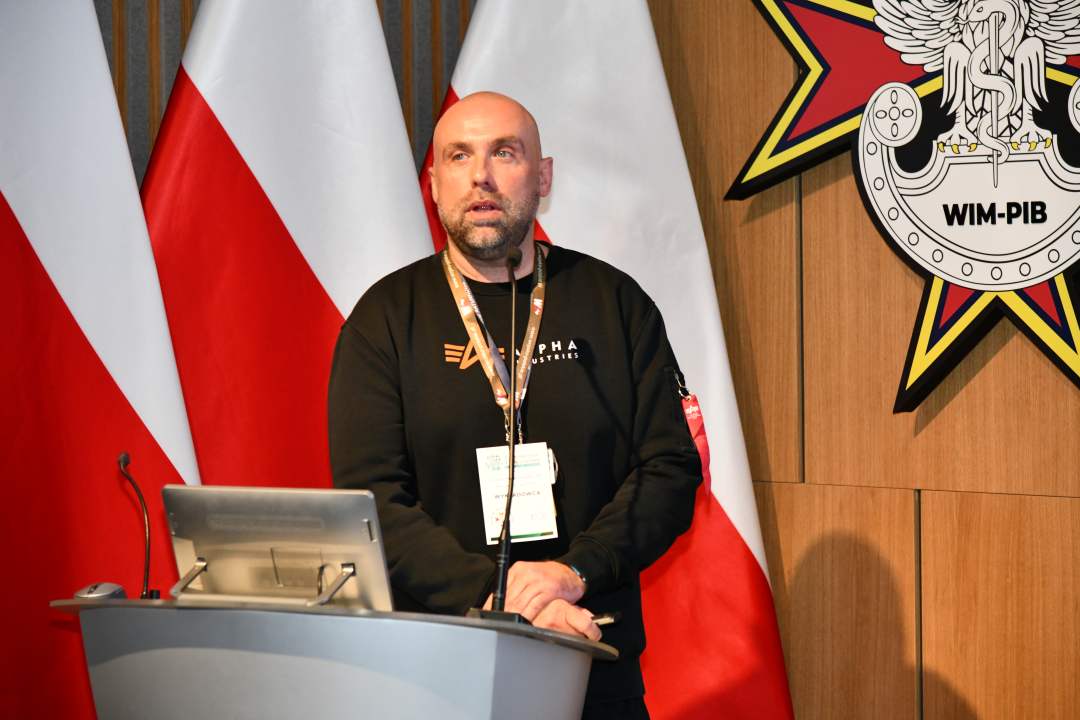 Paweł Witt przy mównicy w sali Wojskowego Instytutu Medycznego; za nim trzy polskie flagi i emblemat WIM‑PIB.