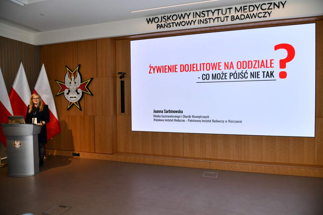 Joanna Sarbinowska stoi przy mównicy w sali Wojskowego Instytutu Medycznego; za nią trzy polskie flagi i emblemat WIM‑PIB. Na ekranie widoczny slajd z tytułem prezentacji ‘Żywienie dojelitowe na oddziale – co może pójść nie tak?