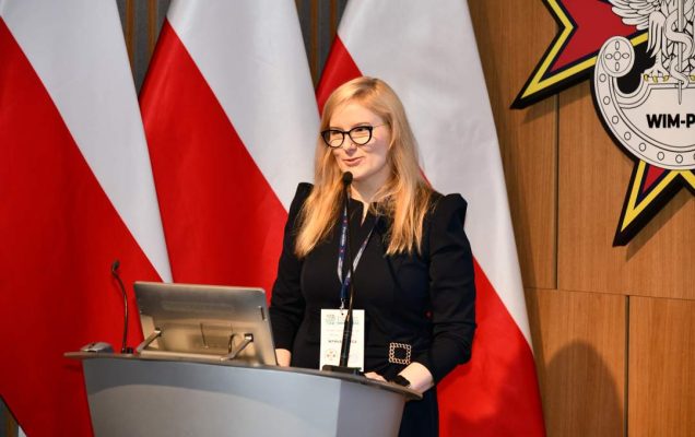 Joanna Sarbinowska stoi przy mównicy w sali Wojskowego Instytutu Medycznego; za nią trzy polskie flagi i emblemat WIM‑PIB.