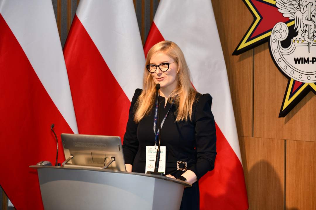 Joanna Sarbinowska stoi przy mównicy w sali Wojskowego Instytutu Medycznego; za nią trzy polskie flagi i emblemat WIM‑PIB.