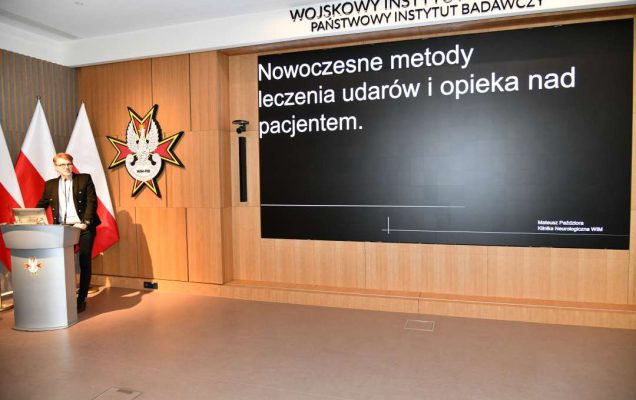 Mateusz Paździora przy mównicy w sali Wojskowego Instytutu Medycznego; za nim dwie polskie flagi i emblemat WIM‑PIB. Na ekranie widoczny slajd z tytułem prezentacji ‘Nowoczesne metody leczenia udarów i opieka nad pacjentem