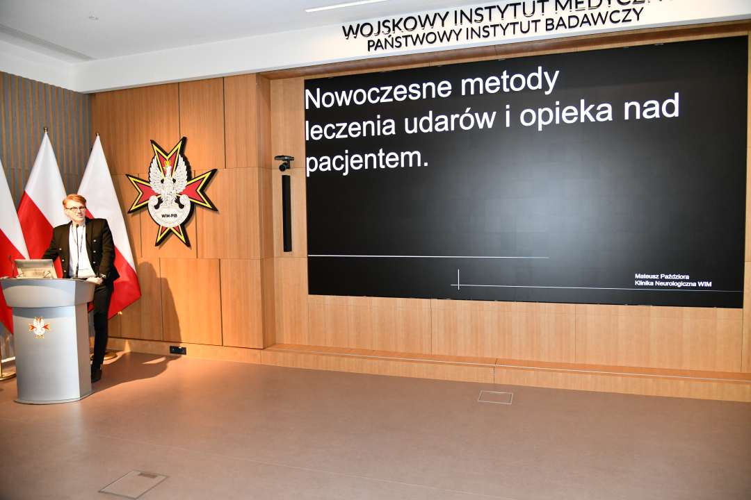 Mateusz Paździora przy mównicy w sali Wojskowego Instytutu Medycznego; za nim dwie polskie flagi i emblemat WIM‑PIB. Na ekranie widoczny slajd z tytułem prezentacji ‘Nowoczesne metody leczenia udarów i opieka nad pacjentem