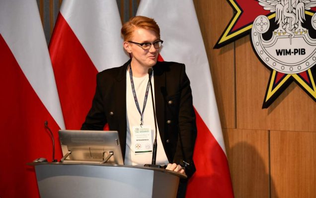 Mateusz Paździora przy mównicy w sali Wojskowego Instytutu Medycznego; za nim dwie polskie flagi i emblemat WIM‑PIB.