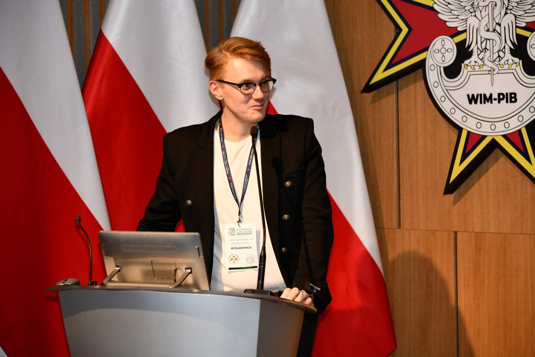 Mateusz Paździora przy mównicy w sali Wojskowego Instytutu Medycznego; za nim dwie polskie flagi i emblemat WIM‑PIB.