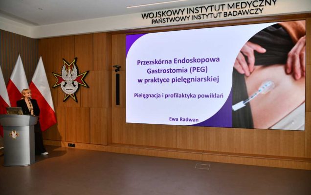 Prelegentka przy mównicy w sali Wojskowego Instytutu Medycznego; obok polska flaga i emblemat WIM‑PIB. Na ekranie widoczny slajd z tytułem ‘Przezskórna endoskopowa gastrostomia (PEG) w praktyce pielęgniarskiej – pielęgnacja i profilaktyka powikłań.