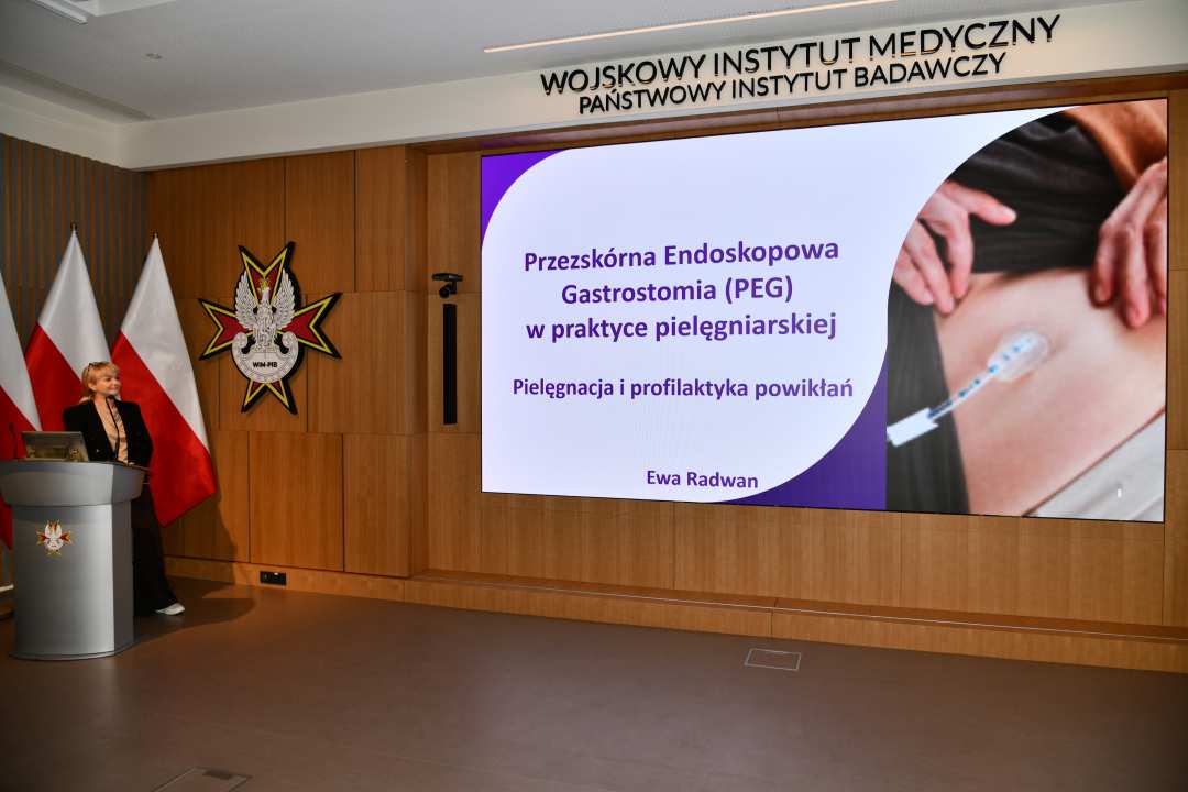 Prelegentka przy mównicy w sali Wojskowego Instytutu Medycznego; obok polska flaga i emblemat WIM‑PIB. Na ekranie widoczny slajd z tytułem ‘Przezskórna endoskopowa gastrostomia (PEG) w praktyce pielęgniarskiej – pielęgnacja i profilaktyka powikłań.