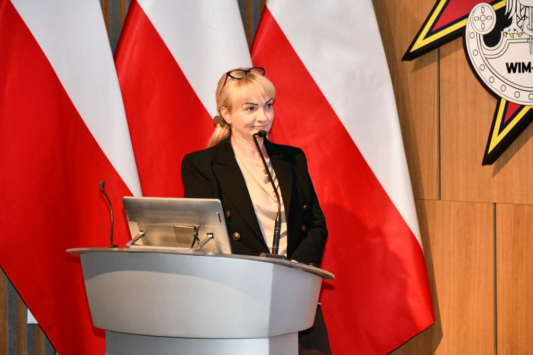 Prelegentka przy mównicy w sali Wojskowego Instytutu Medycznego; obok polska flaga i emblemat WIM‑PIB.