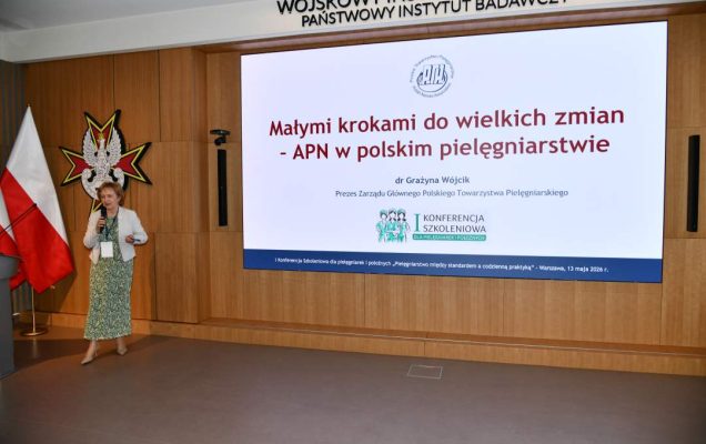 Prelegentka stoi na scenie Wojskowego Instytutu Medycznego; obok dwie polskie flagi i emblemat WIM‑PIB. Na ekranie widoczny slajd z tytułem prezentacji ‘Małymi krokami do wielkich zmian – APN w polskim pielęgniarstwie'.
