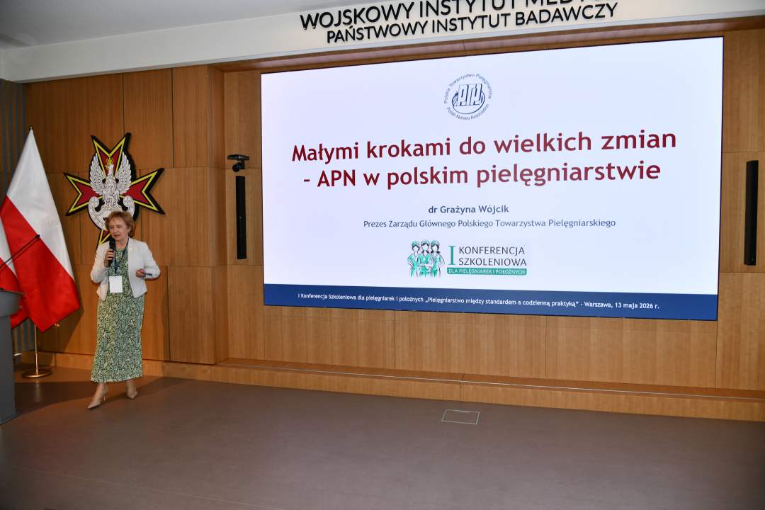 Prelegentka stoi na scenie Wojskowego Instytutu Medycznego; obok dwie polskie flagi i emblemat WIM‑PIB. Na ekranie widoczny slajd z tytułem prezentacji ‘Małymi krokami do wielkich zmian – APN w polskim pielęgniarstwie'.
