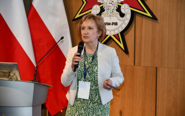 Prelegentka stoi na scenie Wojskowego Instytutu Medycznego; obok dwie polskie flagi i emblemat WIM‑PIB.