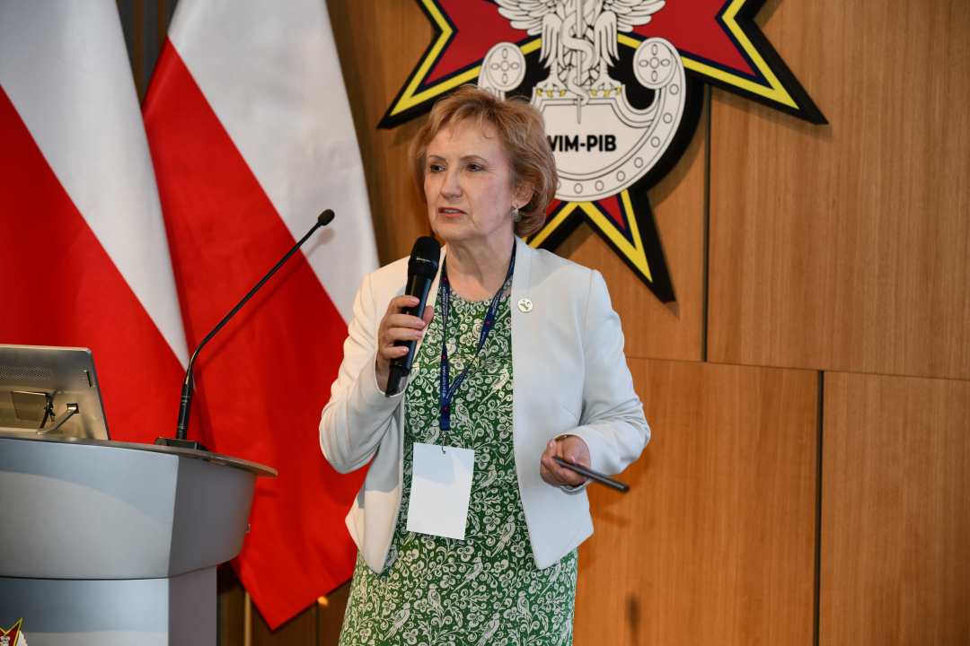 Prelegentka stoi na scenie Wojskowego Instytutu Medycznego; obok dwie polskie flagi i emblemat WIM‑PIB.