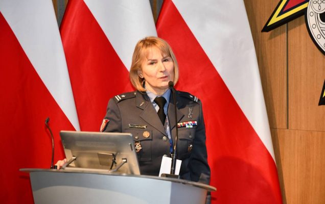Prelegentka w mundurze stoi przy mównicy w sali Wojskowego Instytutu Medycznego; obok polska flaga i emblemat WIM‑PIB.