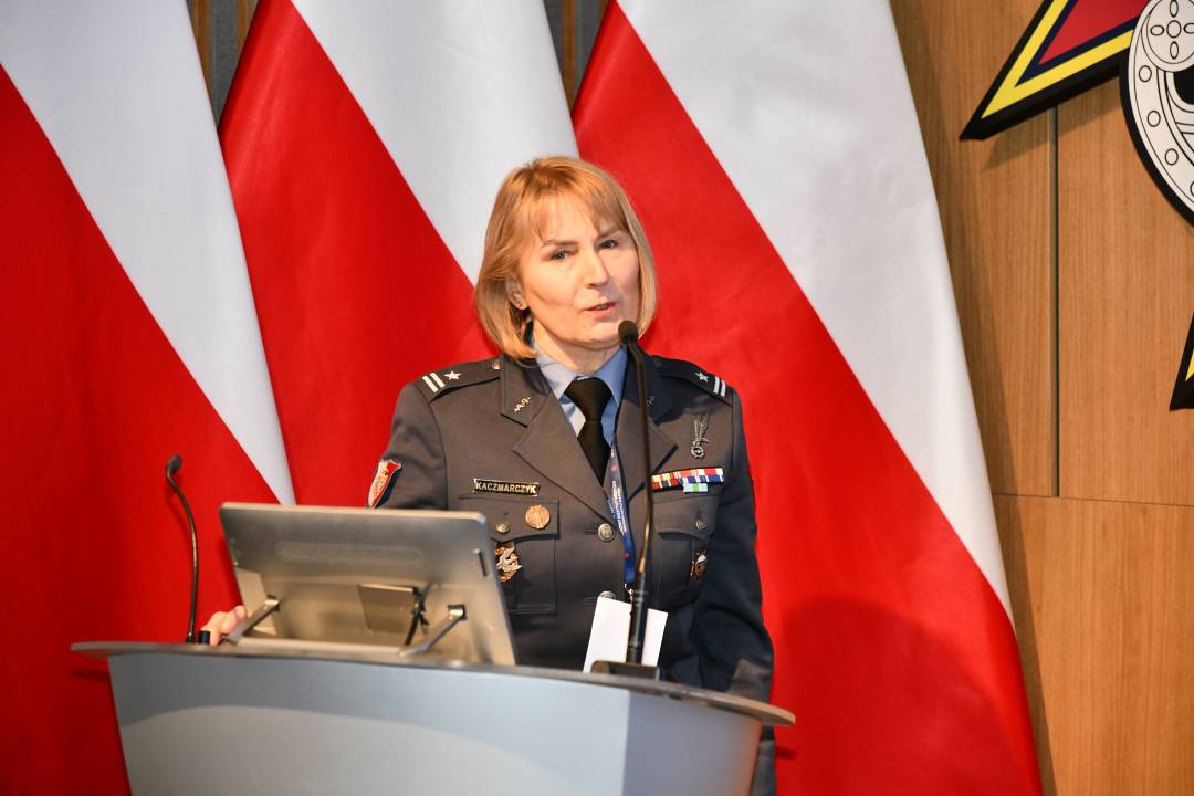 Prelegentka w mundurze stoi przy mównicy w sali Wojskowego Instytutu Medycznego; obok polska flaga i emblemat WIM‑PIB.