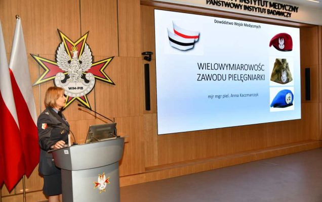 Prelegentka w mundurze stoi przy mównicy w sali Wojskowego Instytutu Medycznego; obok polska flaga i emblemat WIM‑PIB. Na ekranie widoczny slajd z tytułem Wielowymiarowość zawodu pielęgniarki’ oraz grafikami czapek wojskowych.