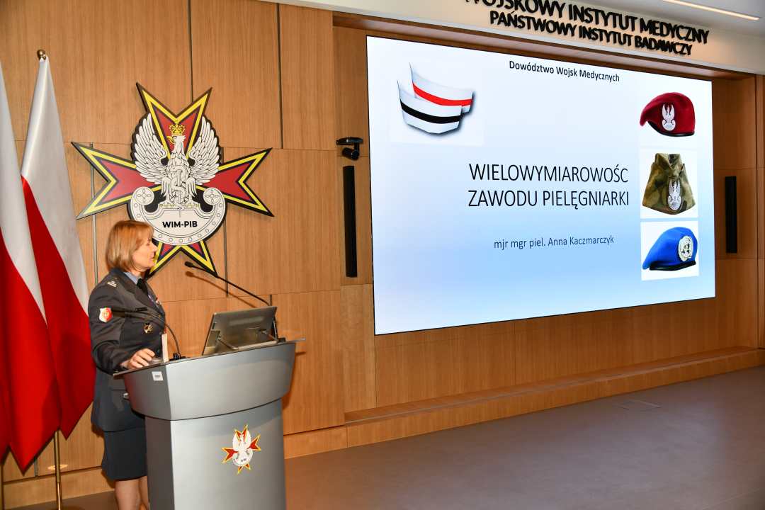 Prelegentka w mundurze stoi przy mównicy w sali Wojskowego Instytutu Medycznego; obok polska flaga i emblemat WIM‑PIB. Na ekranie widoczny slajd z tytułem Wielowymiarowość zawodu pielęgniarki’ oraz grafikami czapek wojskowych.