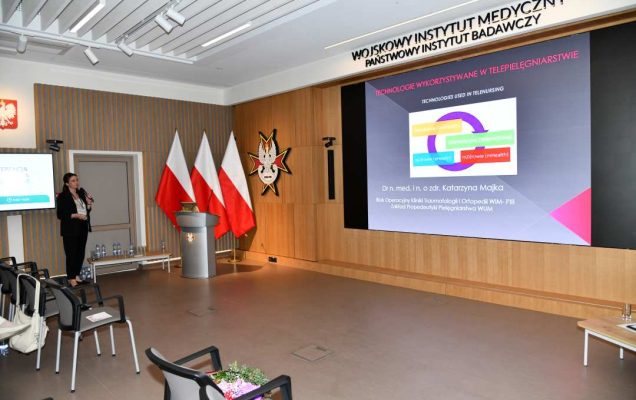 Prelegentka przy mównicy w sali Wojskowego Instytutu Medycznego; obok polskie flagi i emblemat WIM‑PIB. Na ekranie widoczny slajd z tytułem ‘Technologie wykorzystywane w telepielęgniarstwie’ oraz diagramem eHealth i mHealth.