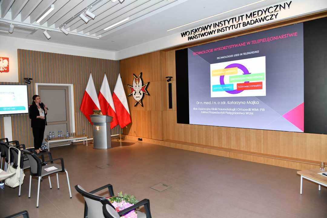 Prelegentka przy mównicy w sali Wojskowego Instytutu Medycznego; obok polskie flagi i emblemat WIM‑PIB. Na ekranie widoczny slajd z tytułem ‘Technologie wykorzystywane w telepielęgniarstwie’ oraz diagramem eHealth i mHealth.