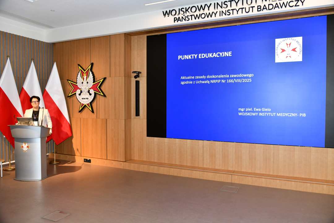 Prelegentka przy mównicy w sali Wojskowego Instytutu Medycznego; za nią trzy polskie flagi i emblemat WIM‑PIB. Na ekranie widoczny slajd z tytułem ‘Punkty edukacyjne – aktualne zasady doskonalenia zawodowego.