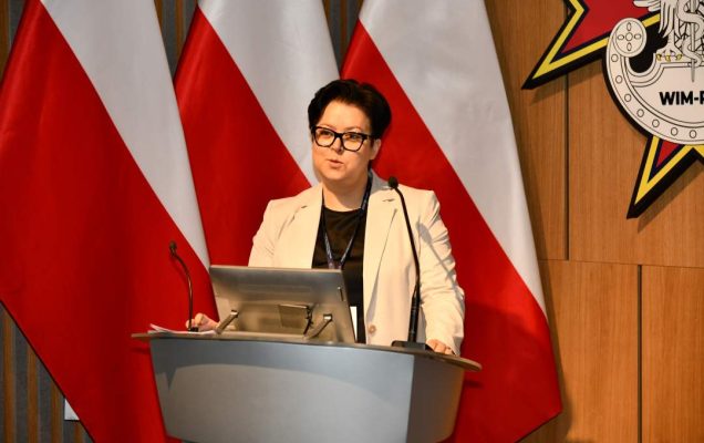 Prelegentka stoi przy mównicy w sali Wojskowego Instytutu Medycznego; obok polska flaga i emblemat WIM‑PIB.