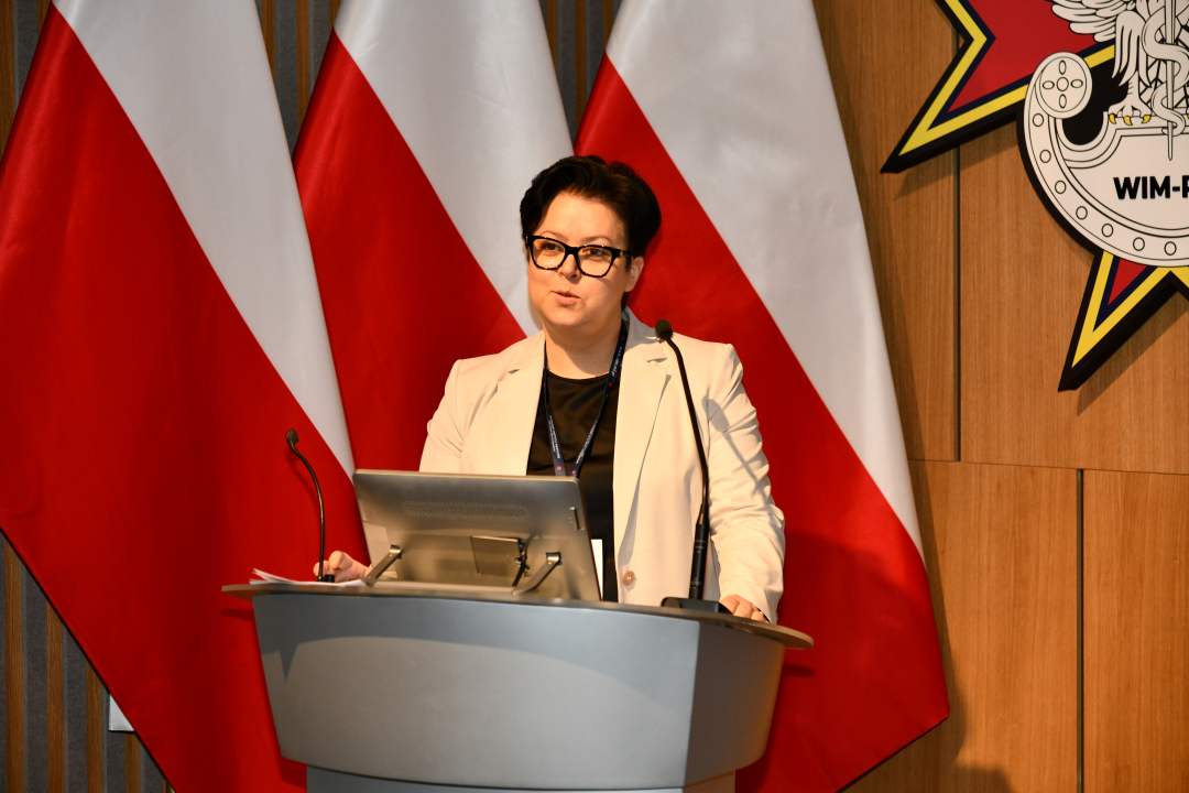 Prelegentka stoi przy mównicy w sali Wojskowego Instytutu Medycznego; obok polska flaga i emblemat WIM‑PIB.
