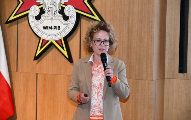 Prelegentka z mikrofonem w ręku w sali Wojskowego Instytutu Medycznego; obok polska flaga i emblemat WIM‑PIB.