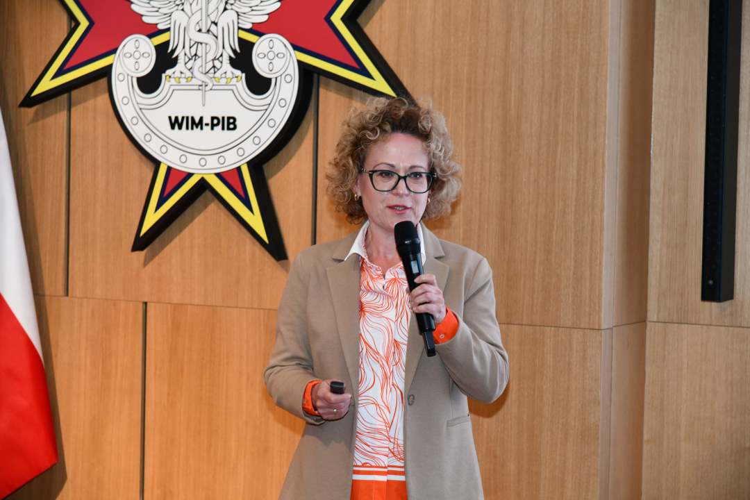 Prelegentka z mikrofonem w ręku w sali Wojskowego Instytutu Medycznego; obok polska flaga i emblemat WIM‑PIB.