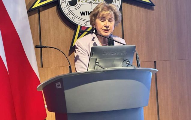 Prelegentka stoi przy mównicy w sali Wojskowego Instytutu Medycznego; obok polska flaga i emblemat WIM‑PIB.
