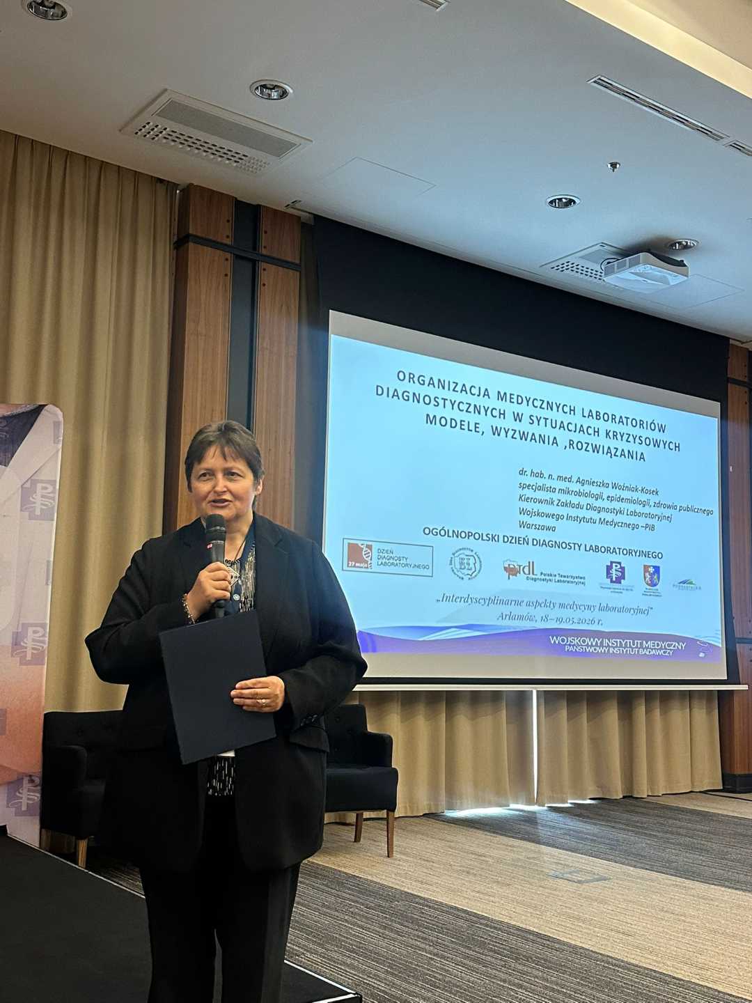 Kierownik Zakładu Diagnostyki Laboratoryjnej dr hab. n. med. Agnieszka Woźniak-Kosek z mikrofonem w ręku na sali konferencyjnej za nią na dużym ekranie wyświetlony slajd z prezentacji.