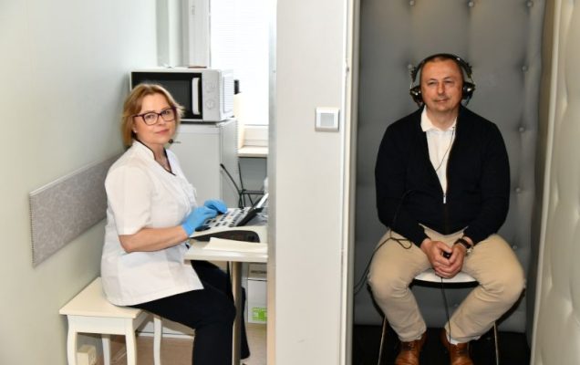 Badanie słuchu w gabinecie laryngologicznym: specjalistka obsługuje audiometr, a pacjent w kabinie ciszy ma nałożone słuchawki.