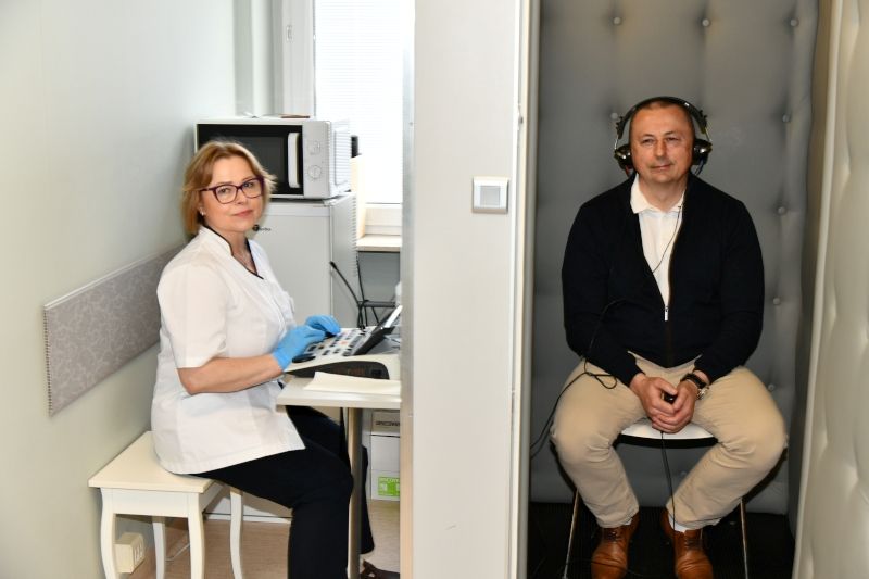 Badanie słuchu w gabinecie laryngologicznym: specjalistka obsługuje audiometr, a pacjent w kabinie ciszy ma nałożone słuchawki.