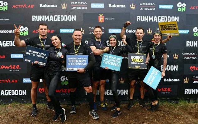 Siedmiu uczestników zawodów Runmageddon stojących razem na tle czarnej ścianki z logotypami sponsorów, m.in. Go On, Activlab, Bakoma, OstroVit, Omoda, Jneeco oraz herbem Warszawy. Wszyscy ubrani są w czarne sportowe stroje z napisem „Runmageddon”, mają medale na szyjach i są ubrudzeni błotem, co wskazuje na ukończenie ekstremalnego biegu terenowego. Uczestnicy uśmiechają się i trzymają kolorowe tabliczki z hasłami, m.in. „Dobre kino się tu odbyło”, „Running because therapy is expensive”, „Nie boję się trudnego terenu”, „Boskie ciało, luźna głowa”, „Forma sprawdzona, bo na białku zrobiona” oraz „Offroad mode: ON”.