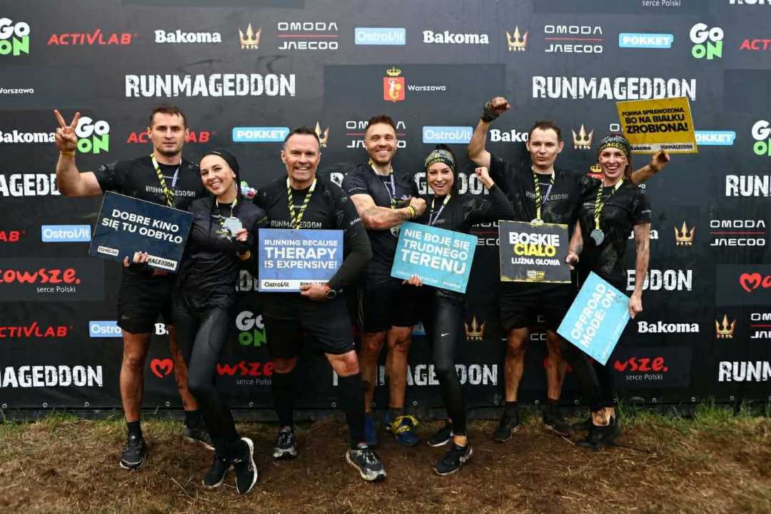 Siedmiu uczestników zawodów Runmageddon stojących razem na tle czarnej ścianki z logotypami sponsorów, m.in. Go On, Activlab, Bakoma, OstroVit, Omoda, Jneeco oraz herbem Warszawy. Wszyscy ubrani są w czarne sportowe stroje z napisem „Runmageddon”, mają medale na szyjach i są ubrudzeni błotem, co wskazuje na ukończenie ekstremalnego biegu terenowego. Uczestnicy uśmiechają się i trzymają kolorowe tabliczki z hasłami, m.in. „Dobre kino się tu odbyło”, „Running because therapy is expensive”, „Nie boję się trudnego terenu”, „Boskie ciało, luźna głowa”, „Forma sprawdzona, bo na białku zrobiona” oraz „Offroad mode: ON”.