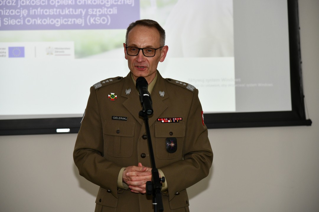 Grzegorz Gielerak przy mikrofonie za nim na dużym ekranie slajd z prezentacji.