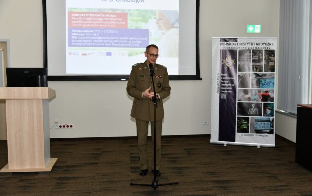 Grzegorz Gielerak podczas przemówienia przy mikrofonie, z anim wyświetlony na dużym ekranie slajd z prezentacji.