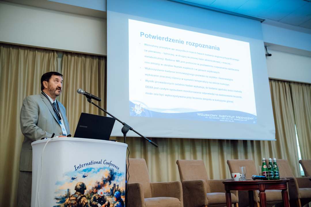 Józef Mróz przy mównicy, za nim ekran z wyświetlonym slajdem prezentacji.