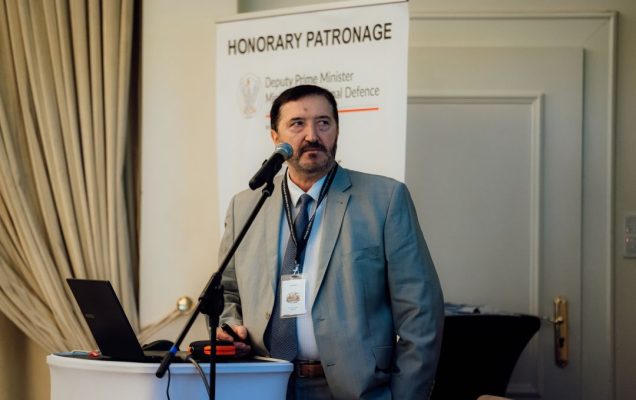 Józef Mróz przy mównicy, za nim plakat konferencji.