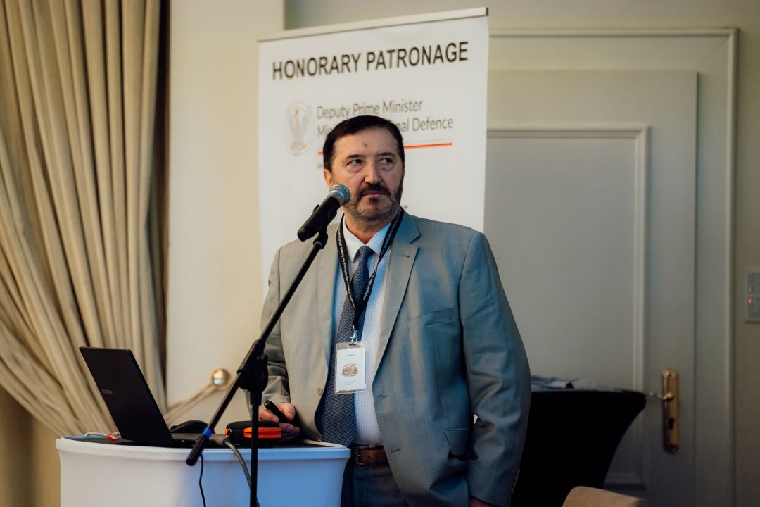 Józef Mróz przy mównicy, za nim plakat konferencji.